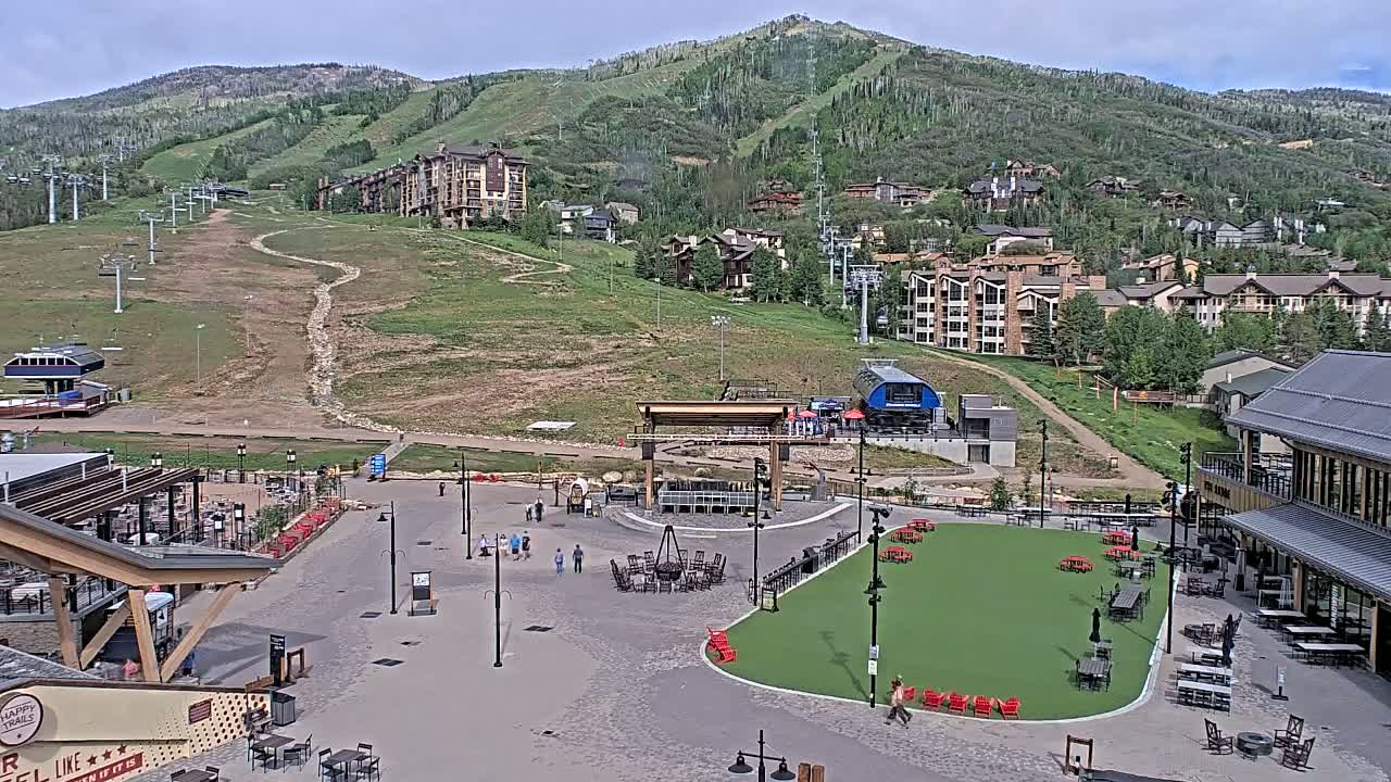 A mountainside village with a paved plaza containing a fire pit, picnic tables, and a small artificial turf area, all under a partly cloudy sky.