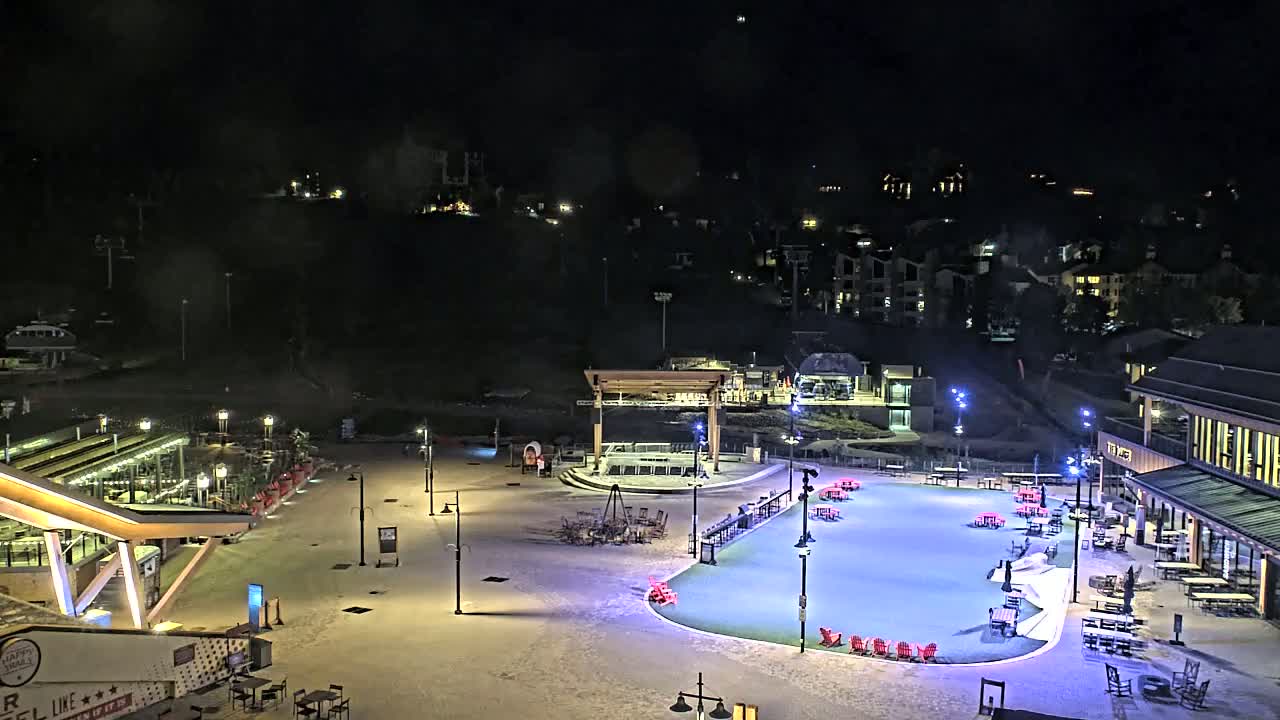 A snow-covered plaza at night, illuminated by lights, features a stage, seating areas, and buildings, with a hillside of houses visible in the background.