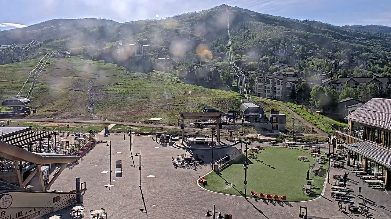 A sunny outdoor scene shows a mountainside with ski lifts, residential buildings, a plaza with outdoor seating areas and a stage, and a grassy area.
