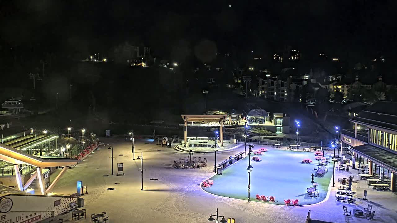 A snow-covered plaza at night, illuminated by lights, features a skating rink, buildings, and outdoor seating areas.