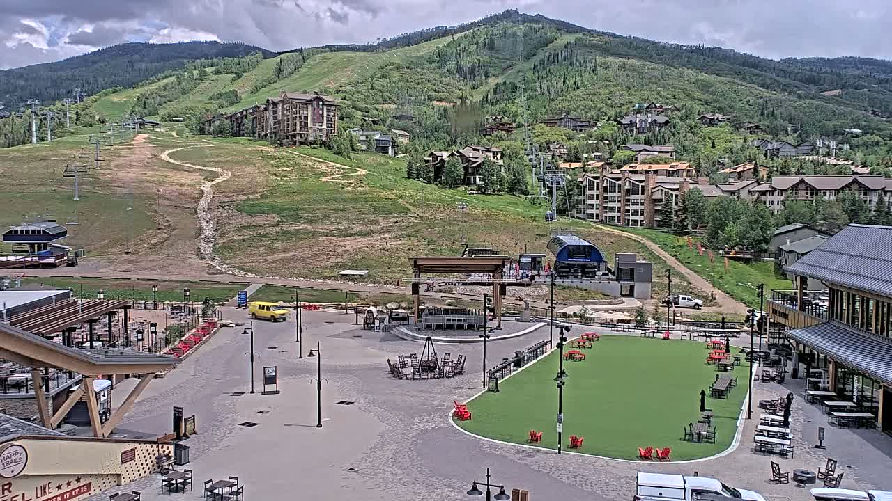 A mountain resort village under partly cloudy skies shows a plaza with outdoor seating areas and a grassy patch, situated below a mountainside with ski lifts and residential buildings.