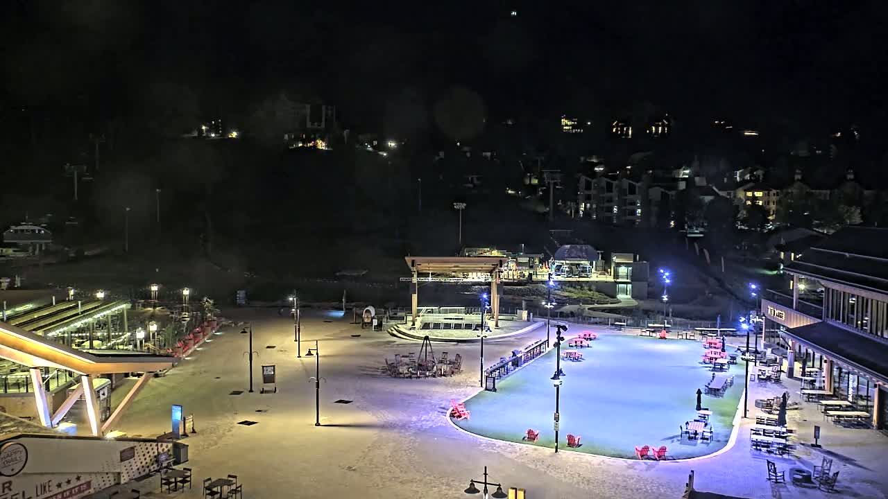 A snow-covered plaza at night with outdoor seating, a stage, and buildings, lit by artificial lights.