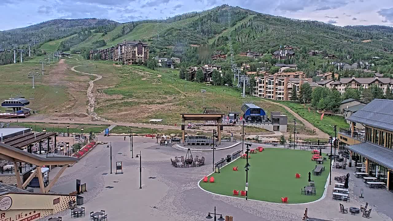 A mountainside resort town under partly cloudy skies features a paved plaza with outdoor seating and a fire pit, overlooking ski lifts and slopes.
