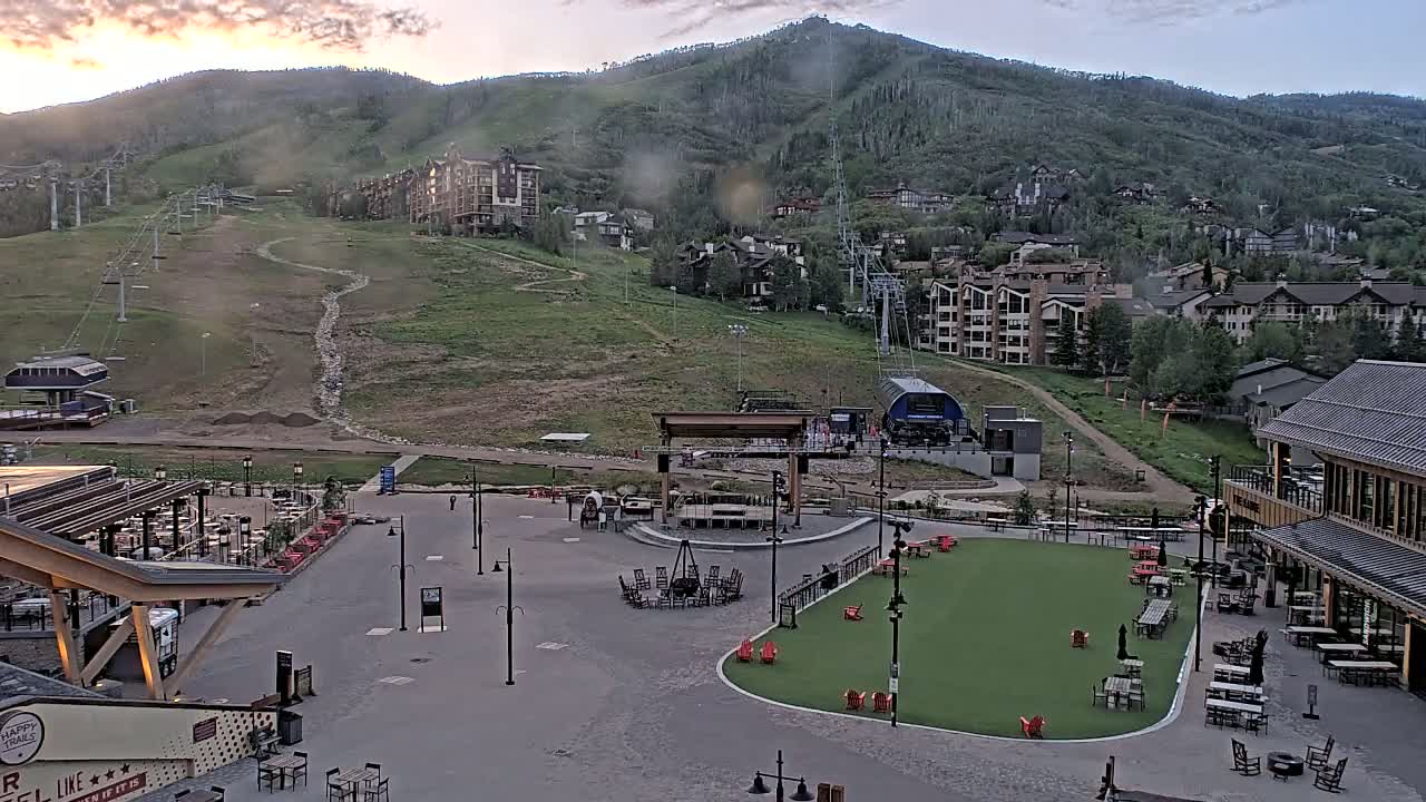 An outdoor plaza with a stage and artificial turf sits below a mountainside with ski lifts and residential buildings, under a partly cloudy, sunrise sky.