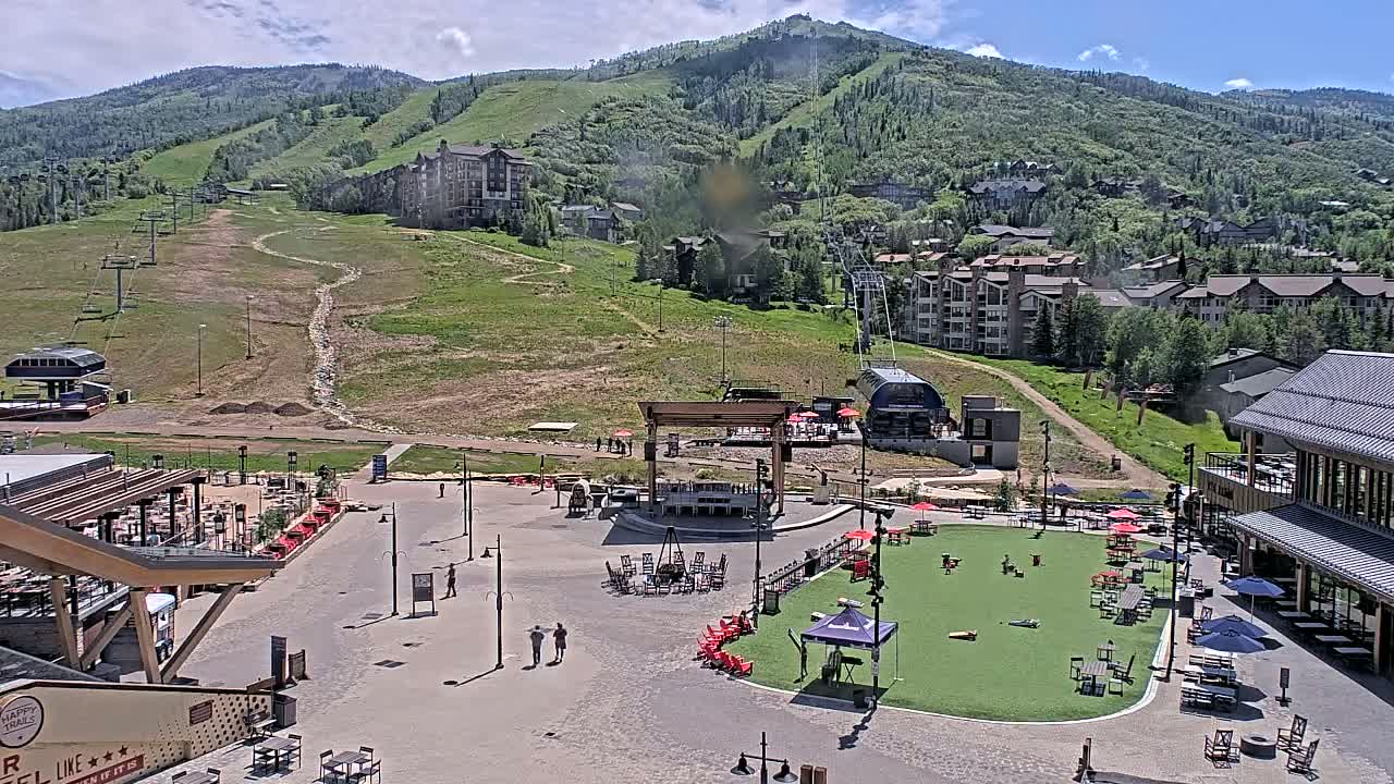 A mountainside village square with a grassy area for games, outdoor seating, and a stage under a partly cloudy summer sky, overlooks ski lifts and slopes on a hillside dotted with buildings.