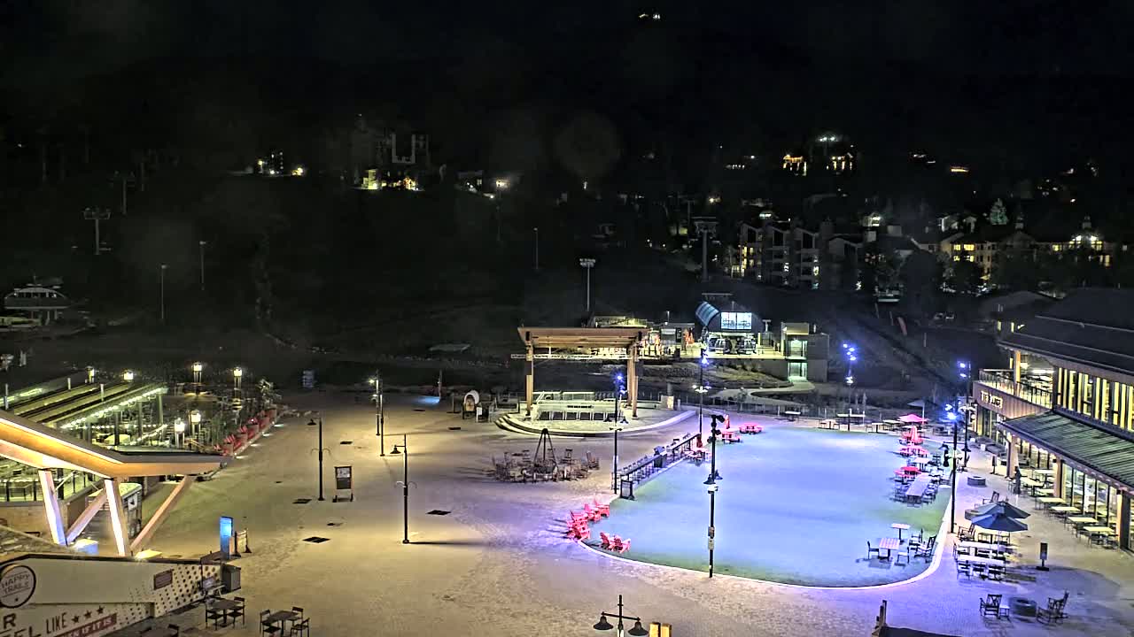 A nighttime aerial view shows a snow-covered plaza with outdoor seating areas, buildings, and a lift for a ski resort, illuminated under artificial light.