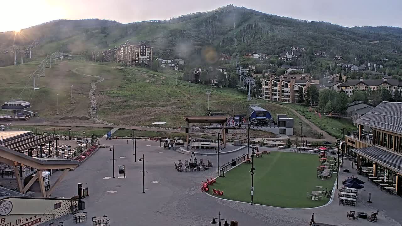 An outdoor plaza with seating areas and a stage is situated at the base of a mountain, with ski lifts visible on the hillside under mostly clear skies.