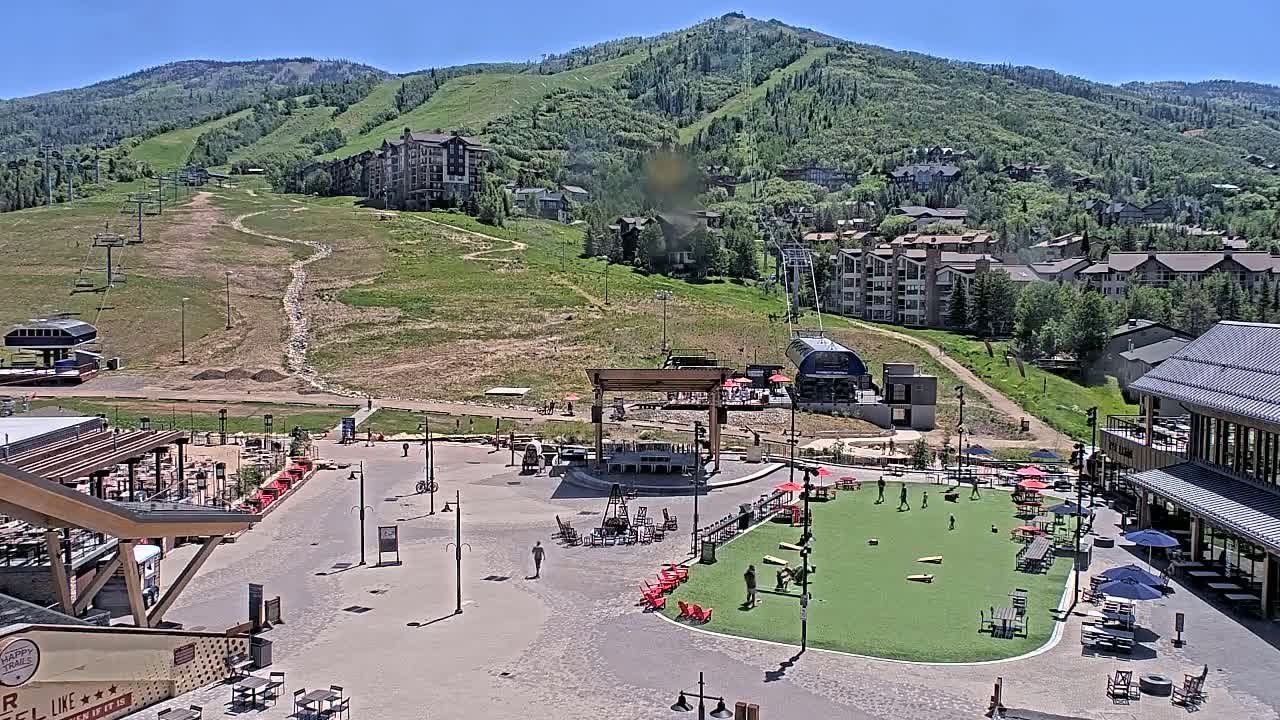 A sunny outdoor scene shows a mountainside village with buildings, grassy areas, an outdoor stage, and people enjoying a plaza with seating and games.