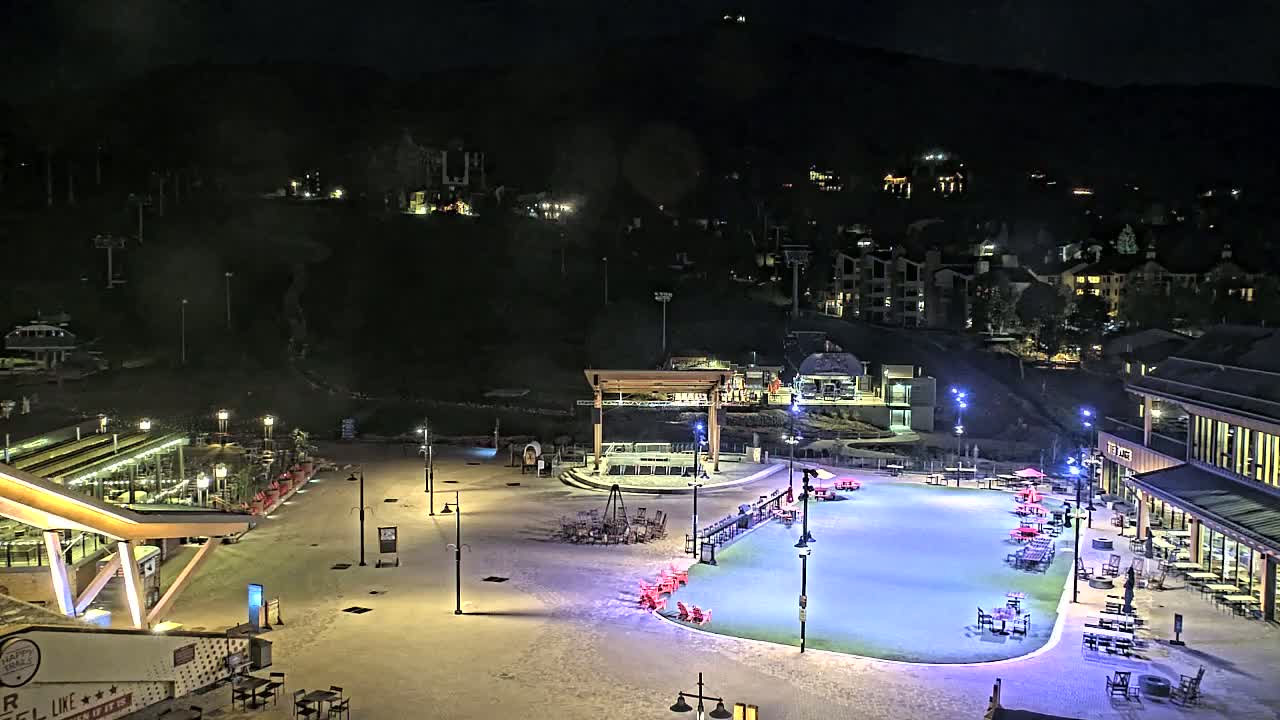 A snow-covered plaza at night, illuminated by lights, features an ice rink, outdoor seating areas, and buildings.