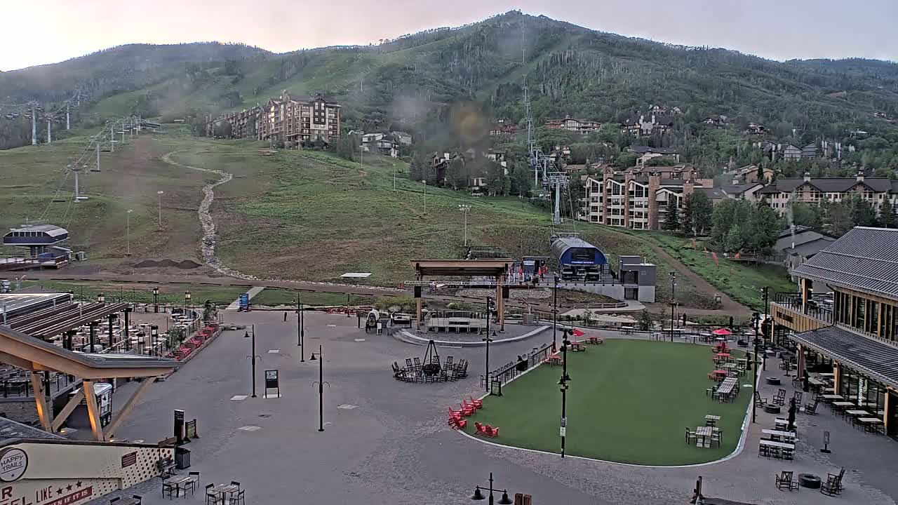A mountainside village with ski lifts, buildings, and an outdoor plaza with seating areas under a partly cloudy sky.