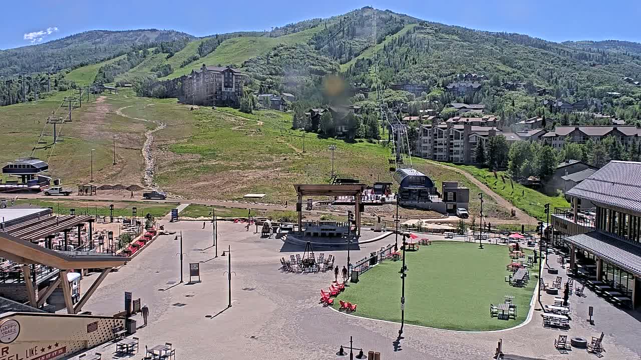 A sunny outdoor scene shows a ski resort base area with a plaza containing a grassy area, an outdoor stage, and numerous tables and chairs, all set against a backdrop of a mountainside with ski lifts and buildings.