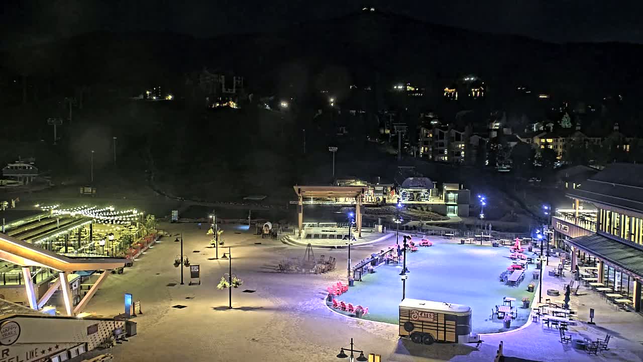 A nighttime view of a snow-covered plaza with outdoor seating, a stage, and an ice rink, surrounded by buildings and a mountain in the background under a clear night sky.