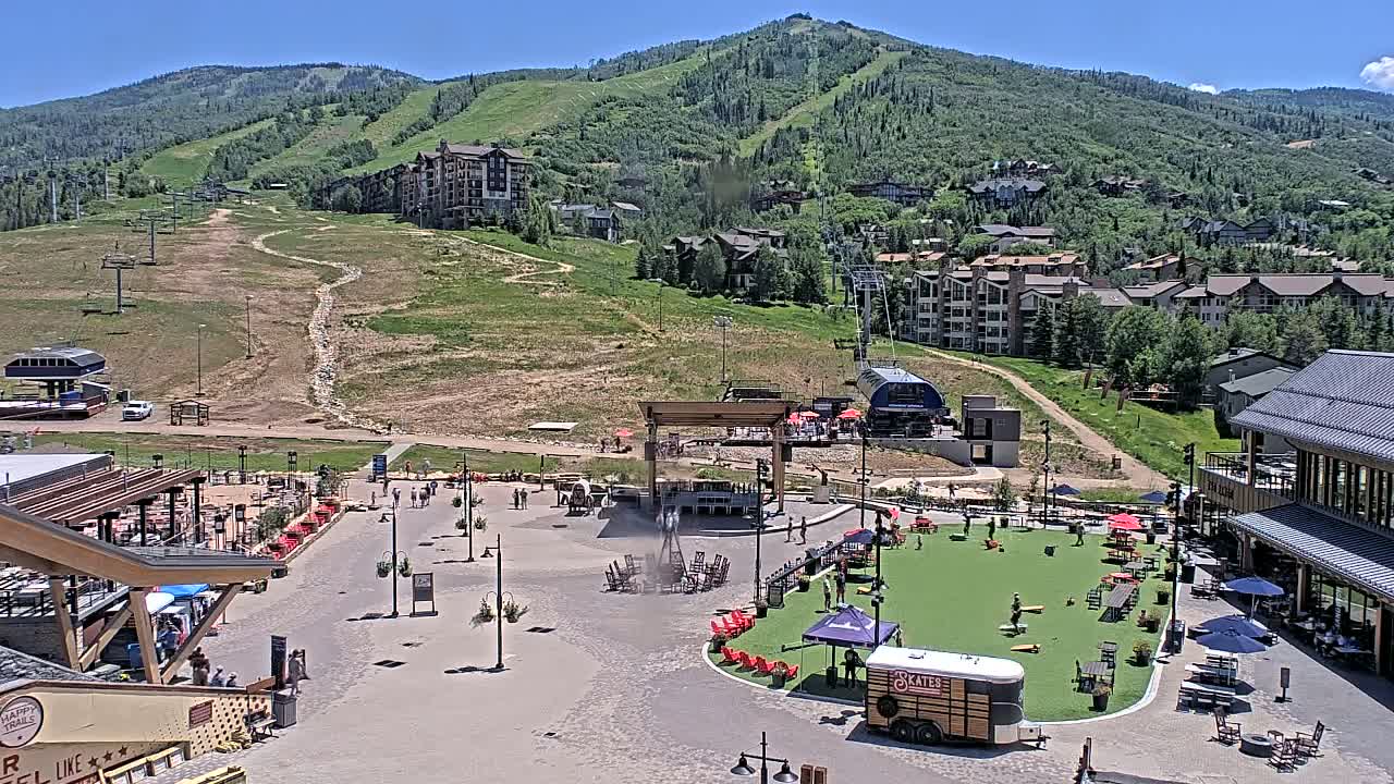 An outdoor plaza with a stage and seating areas is situated at the base of a mountain on a sunny day, with several buildings and ski lifts visible in the background.
