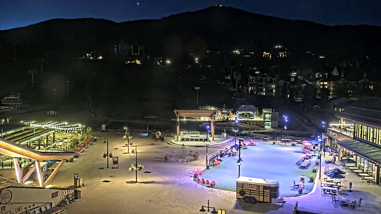 An outdoor ice skating rink and surrounding plaza are illuminated at night, nestled at the base of a dark mountain under a clear sky.