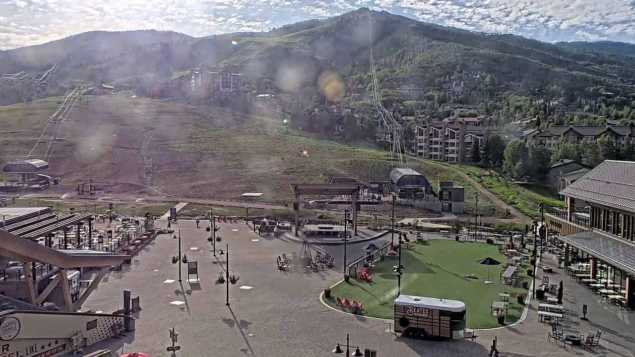 A mostly sunny mountainside village with a ski lift and outdoor plaza containing seating areas and a food truck.