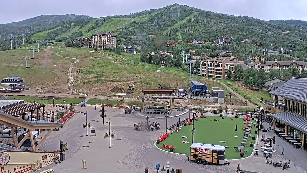 An outdoor plaza with seating areas, a fire pit, and a small artificial turf field sits below a ski resort in a mountain setting under overcast skies.