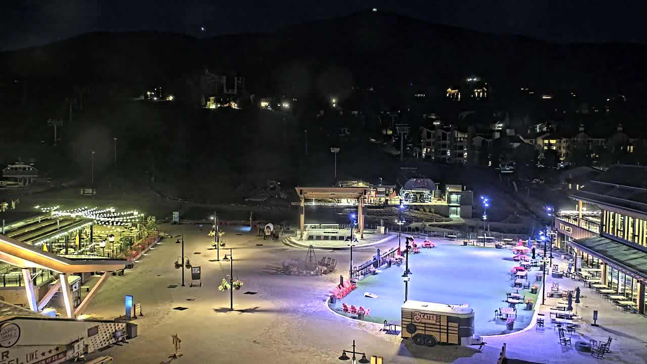 A nighttime, snow-covered plaza with outdoor seating, a skating rink, and buildings illuminated against a dark mountain backdrop.