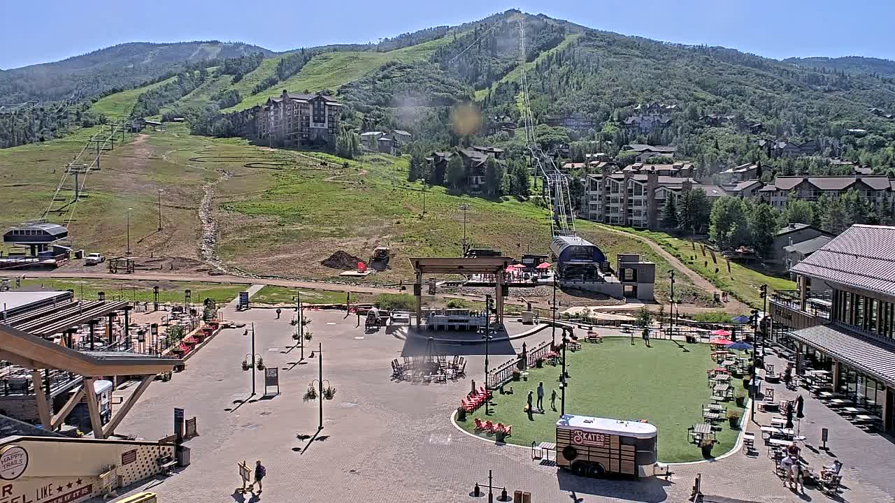 A sunny outdoor scene shows a ski resort base area with buildings, a plaza with seating areas and a food truck, and a mountain with ski lifts in the background.