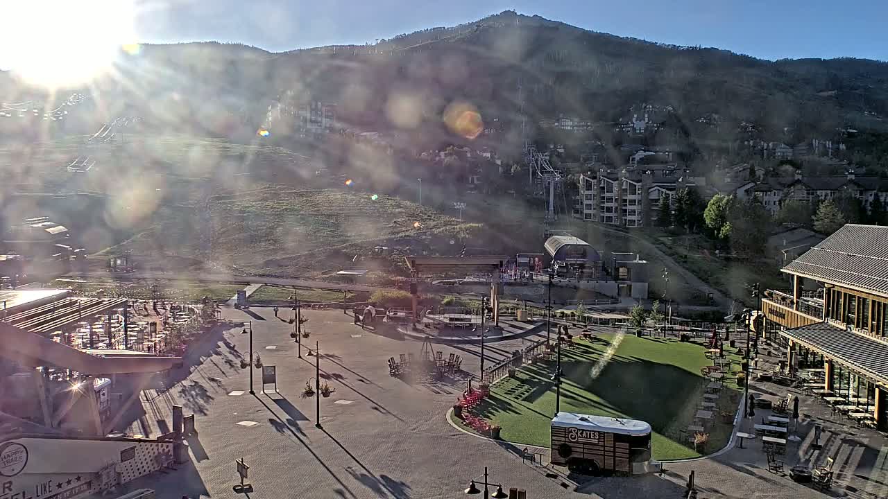 A sunny, hazy morning view of a mountain resort town with outdoor seating areas, a grassy patch, and a ski lift visible on a hillside.
