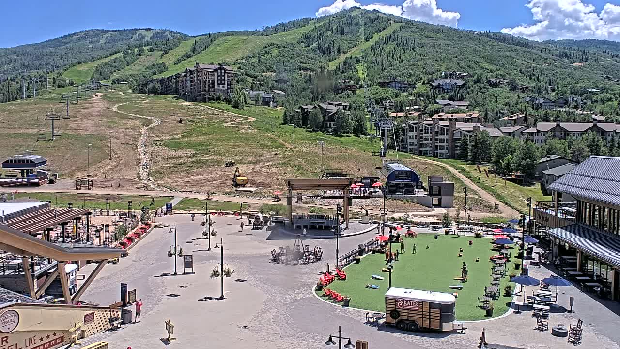 A sunny day reveals a mountainside resort town with a plaza featuring outdoor games and seating areas, all situated below a ski slope with visible lifts and buildings.