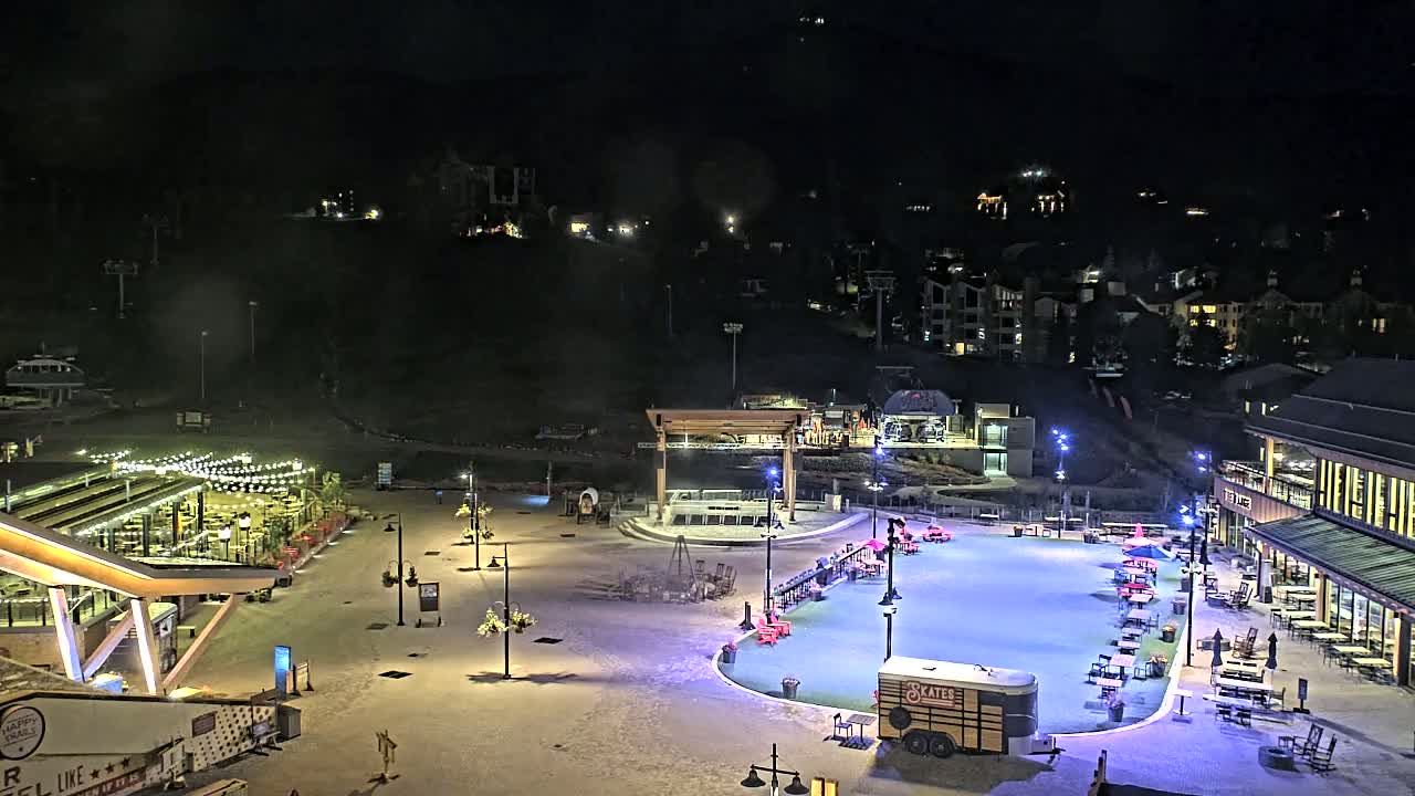 An outdoor nighttime scene shows a plaza with a lighted ice rink, surrounding seating areas, buildings, and a ski lift in the background.