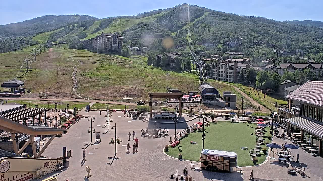 A sunny day reveals a mountainside ski resort village with a plaza featuring outdoor seating, a stage, and a few people walking around.