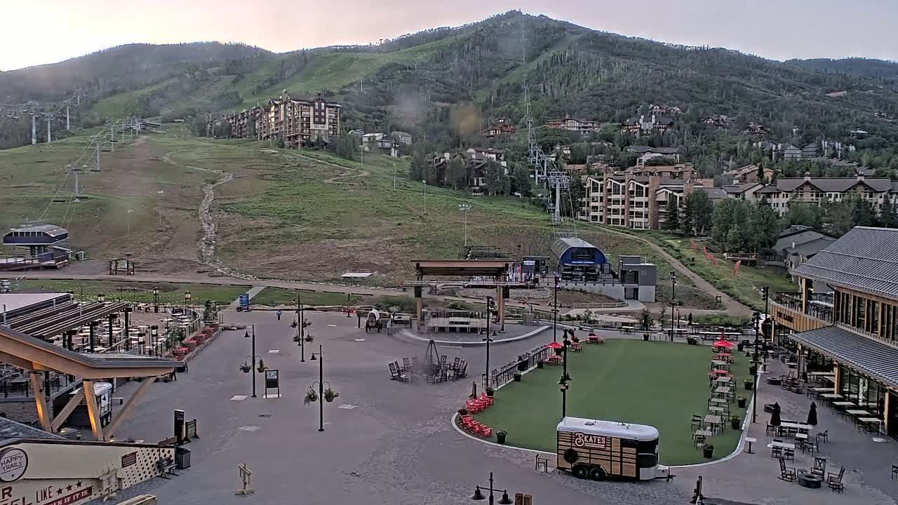 A mountainside village with ski lifts and buildings, a paved plaza with outdoor seating and a fire pit, and a small food trailer under an overcast sky.