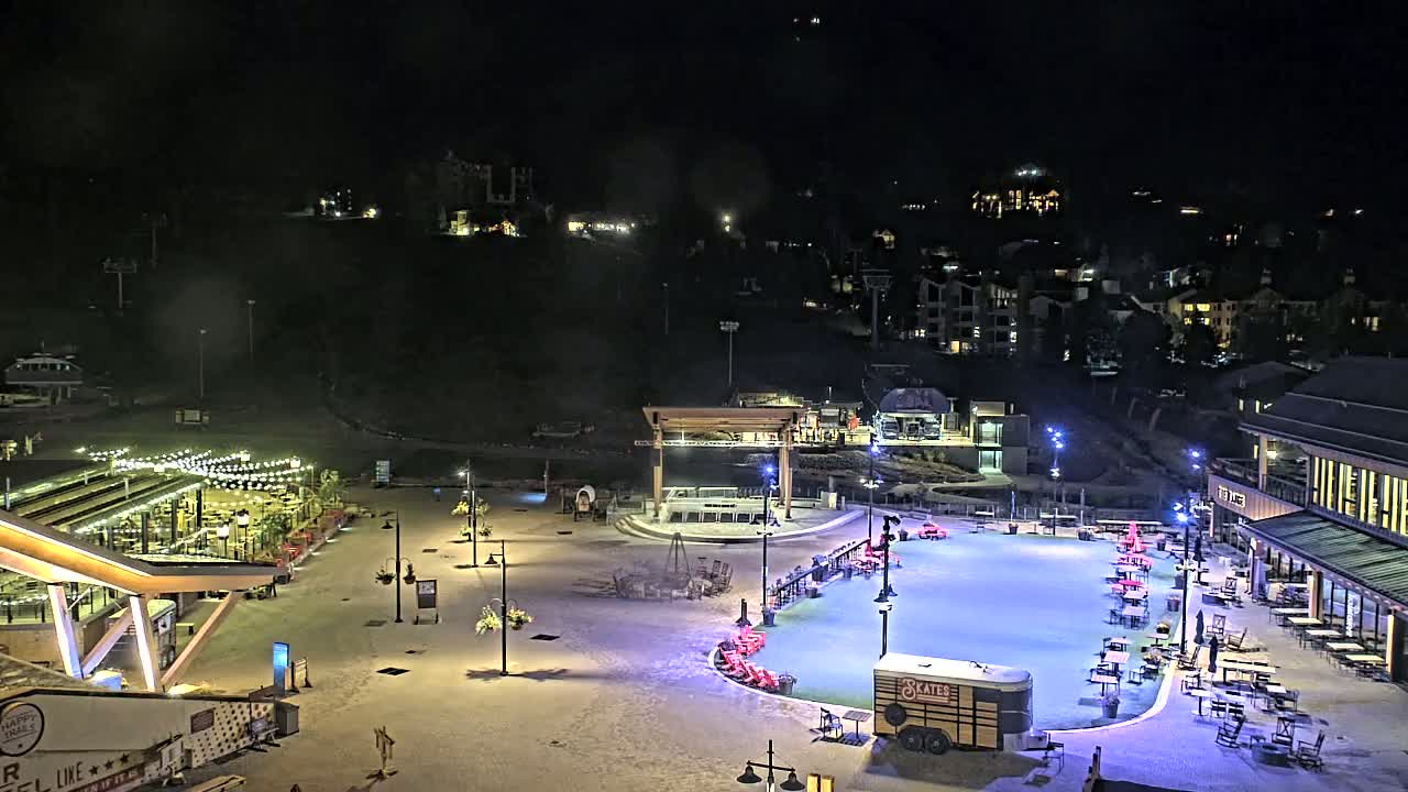 An outdoor ice skating rink is illuminated at night, surrounded by buildings, seating areas, and landscaping, with a light dusting of snow on the ground.