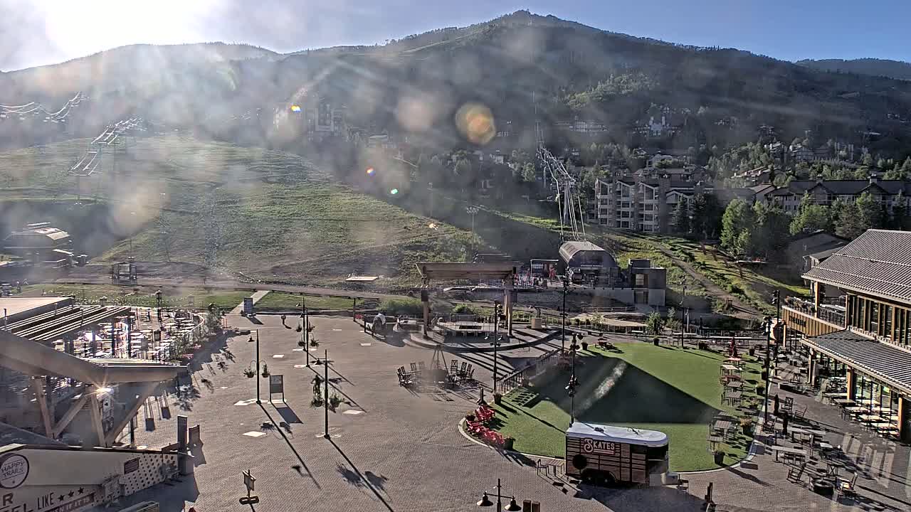 A hazy sunny day reveals a mountainside resort village with a plaza containing outdoor seating and a small patch of grass, all overlooking a ski lift and mountain slope.