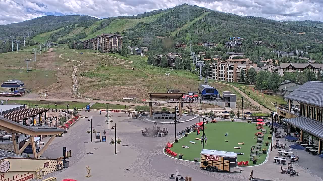 A mountainside village square with a grassy area for games, surrounded by buildings and ski lifts under a partly cloudy sky.