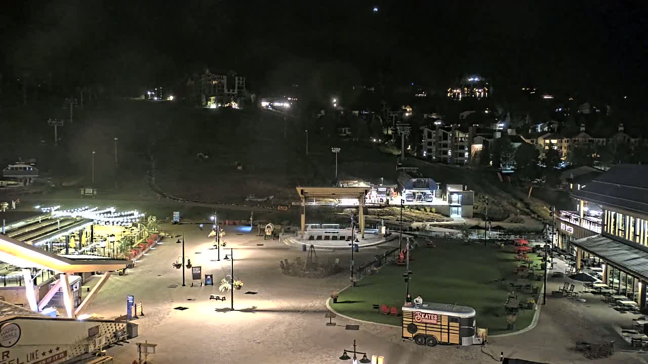 A nighttime aerial view of a village with a plaza featuring outdoor seating, a stage, and a food trailer, all illuminated, overlooking a ski slope and residential buildings on a hill.