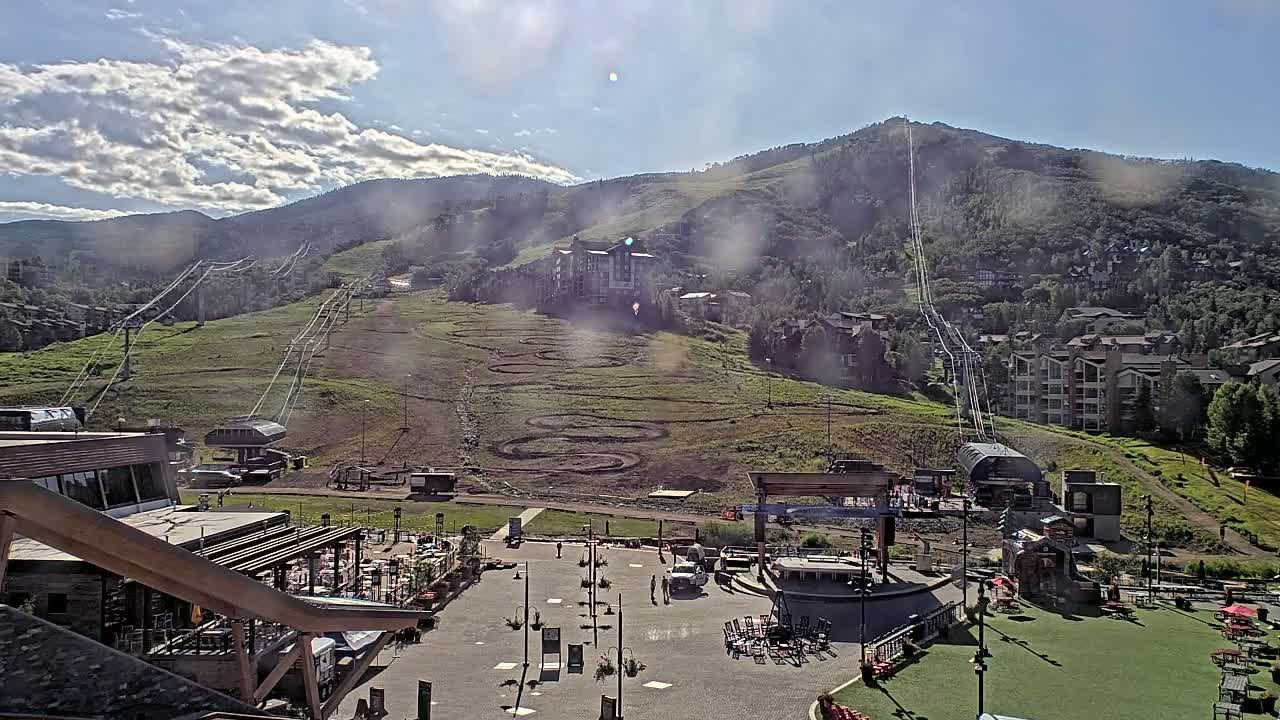 A mostly sunny day reveals a mountainside ski resort with chairlifts, a plaza below, and buildings nestled into the hillside.