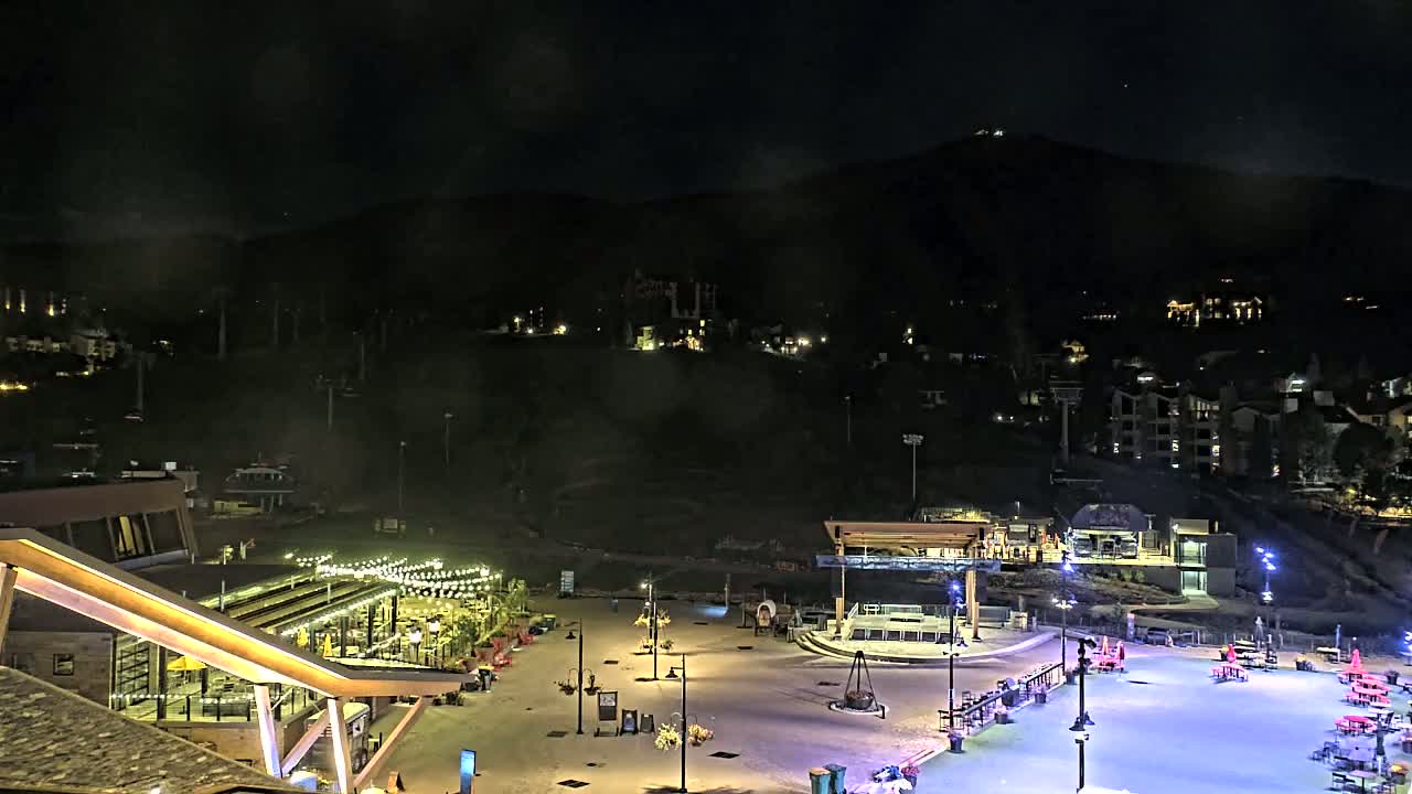 A nighttime view of a snow-covered plaza with buildings and ski lifts on a mountainside under a dark, clear sky.
