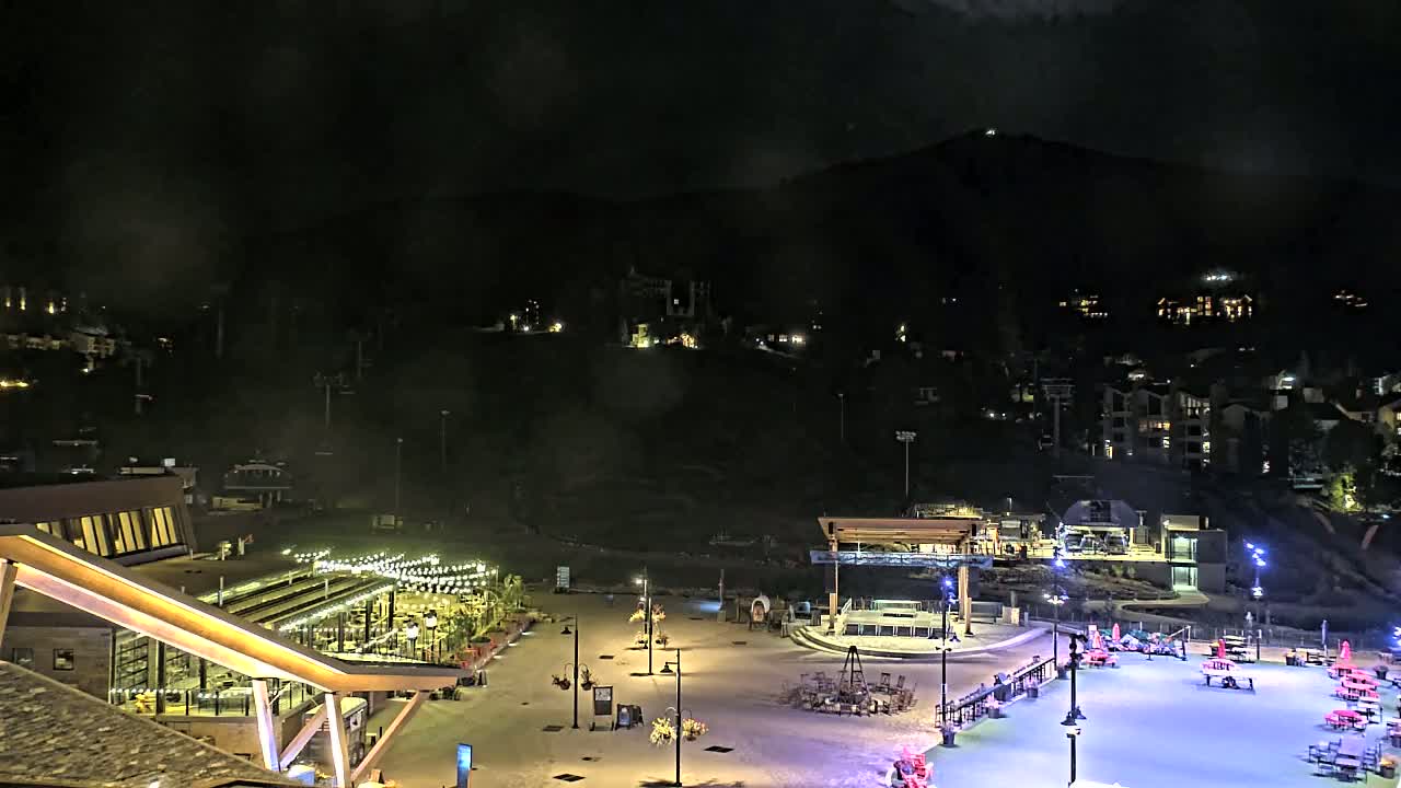 A nighttime view of an illuminated plaza with buildings and a mountain in the background under a cloudy sky.