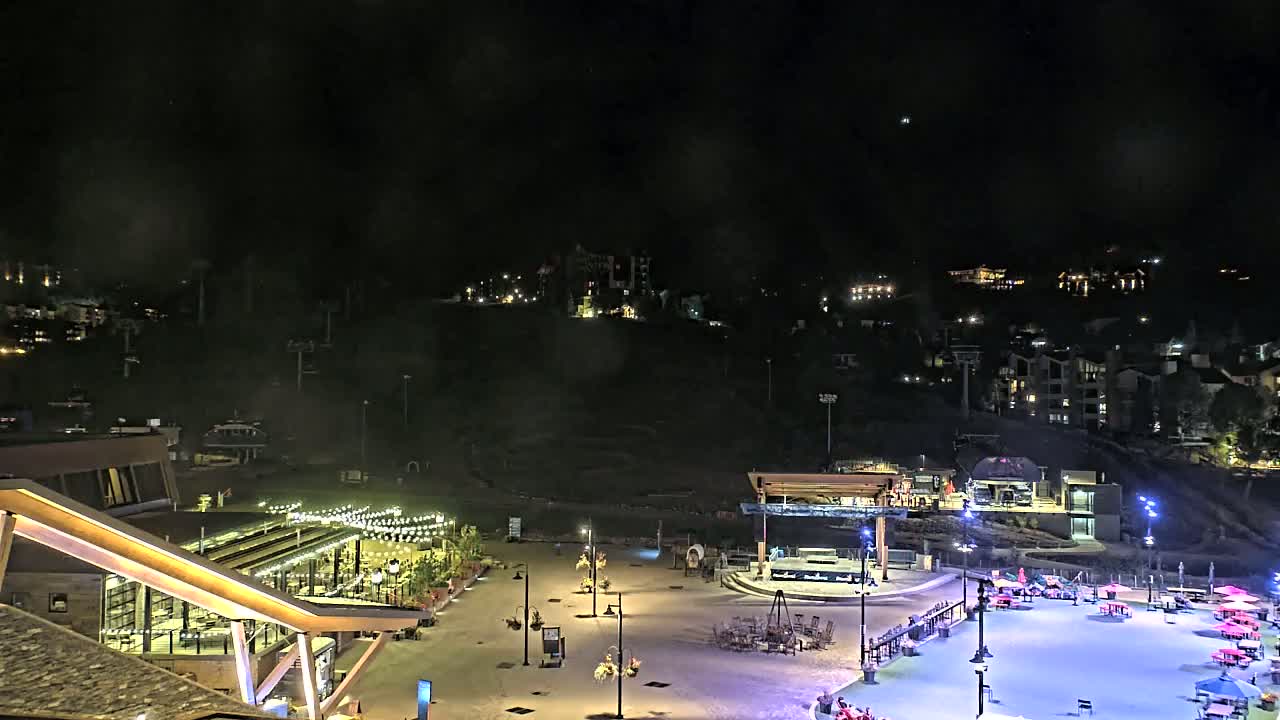 A nighttime view of an illuminated plaza with buildings and a mountain in the background under a cloudy sky.