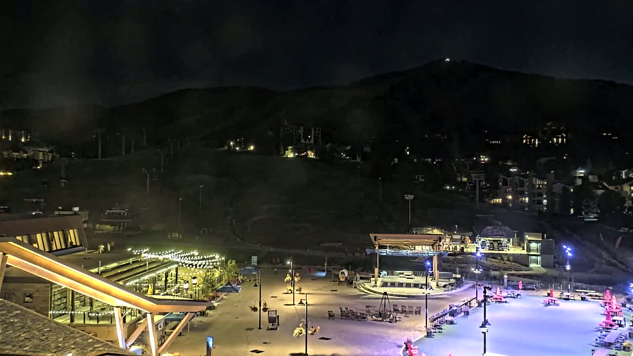A nighttime view of a snow-covered plaza with buildings and lights, situated at the base of a dark mountain with homes and ski lifts visible on its slopes under a hazy sky.