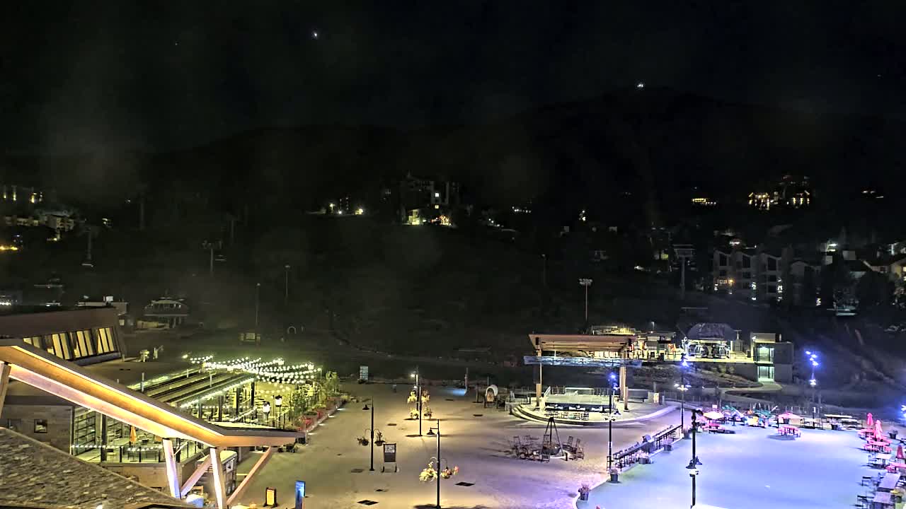 A nighttime view of a snow-covered plaza with buildings and lights, situated at the base of a dark mountain with homes and ski lifts visible on its slopes under a hazy sky.