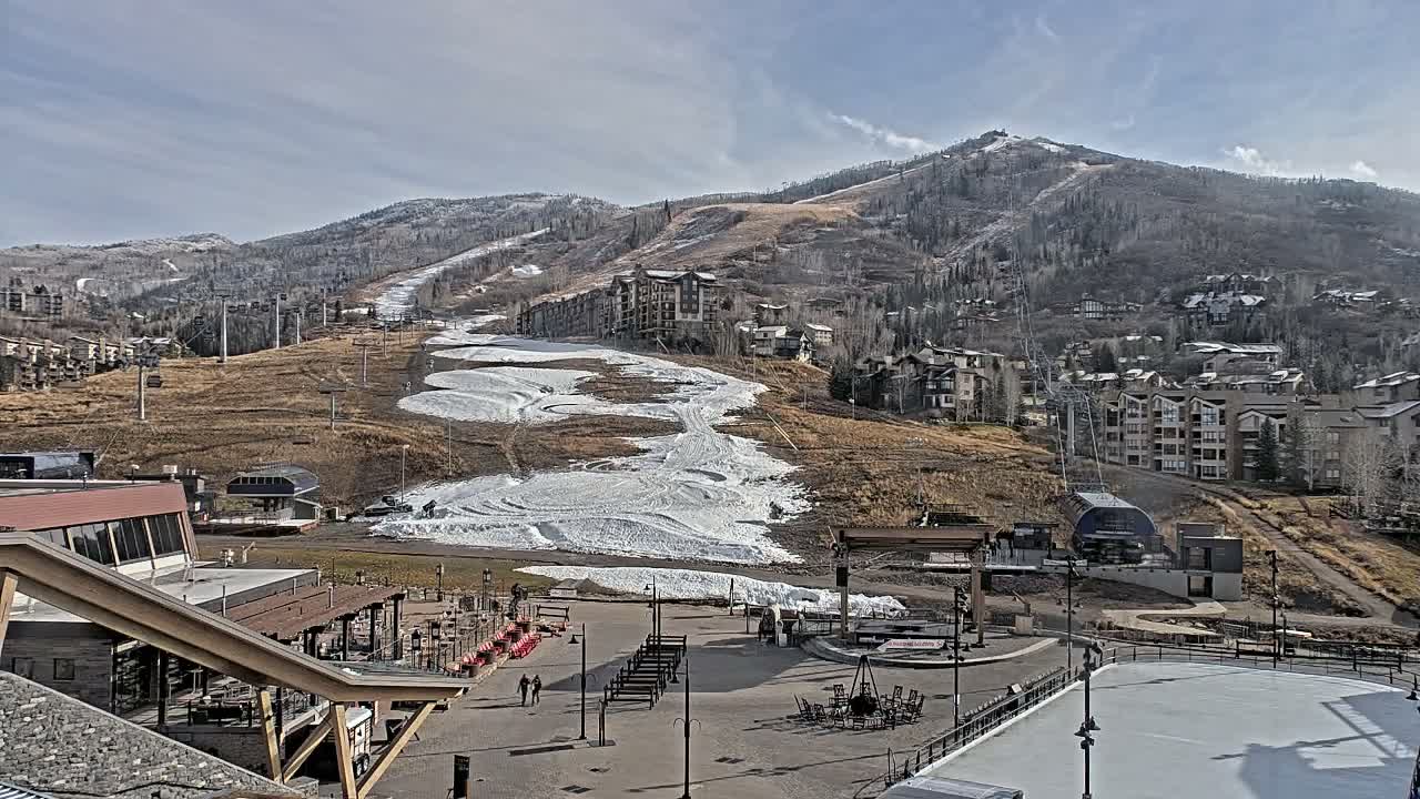 A nighttime view of a snow-covered plaza with buildings and lights, situated at the base of a dark mountain with homes and ski lifts visible on its slopes under a hazy sky.