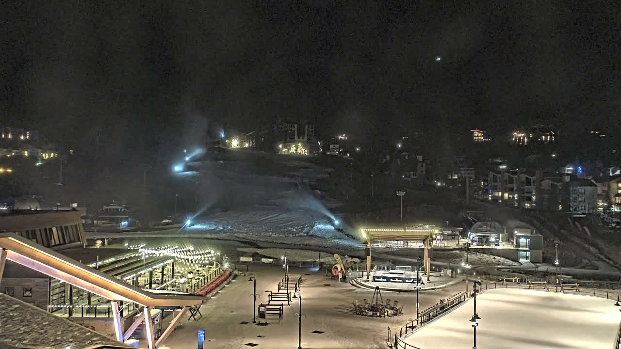 The image shows a brightly illuminated ski resort village at night, featuring a snow-covered mountain slope with active snowmaking, an outdoor dining area, a ski lift, and an ice rink, all under a dark, clear, and cold sky.
