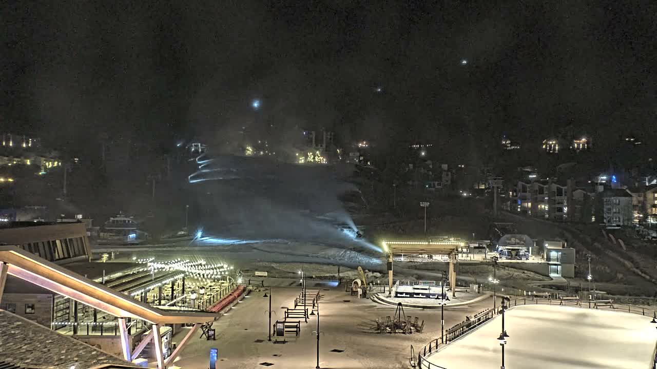 A night view of a ski resort village features active snowmaking on a snowy slope, an illuminated outdoor ice rink, and various buildings with lights under a clear, dark sky.