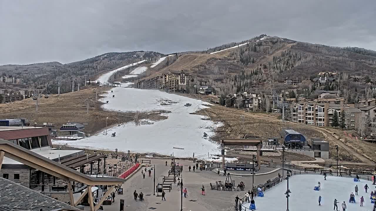 On an overcast day, a bustling mountain ski resort features snow-covered slopes, ski lifts, hillside buildings, a village plaza with pedestrians, and an active outdoor ice skating rink.
