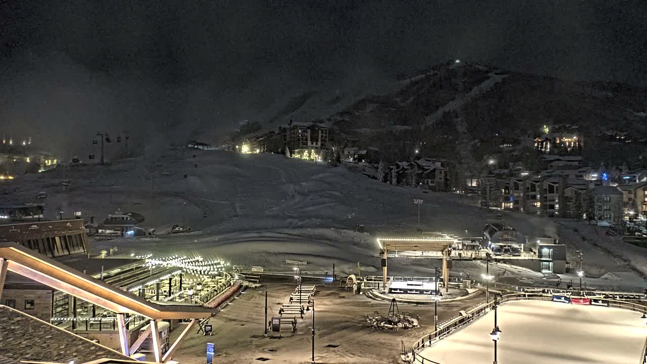 A brightly lit, snow-covered mountain ski resort village is captured at night, showcasing illuminated buildings, ski slopes with lifts, an outdoor dining area, and an ice rink on a cold, dark evening.