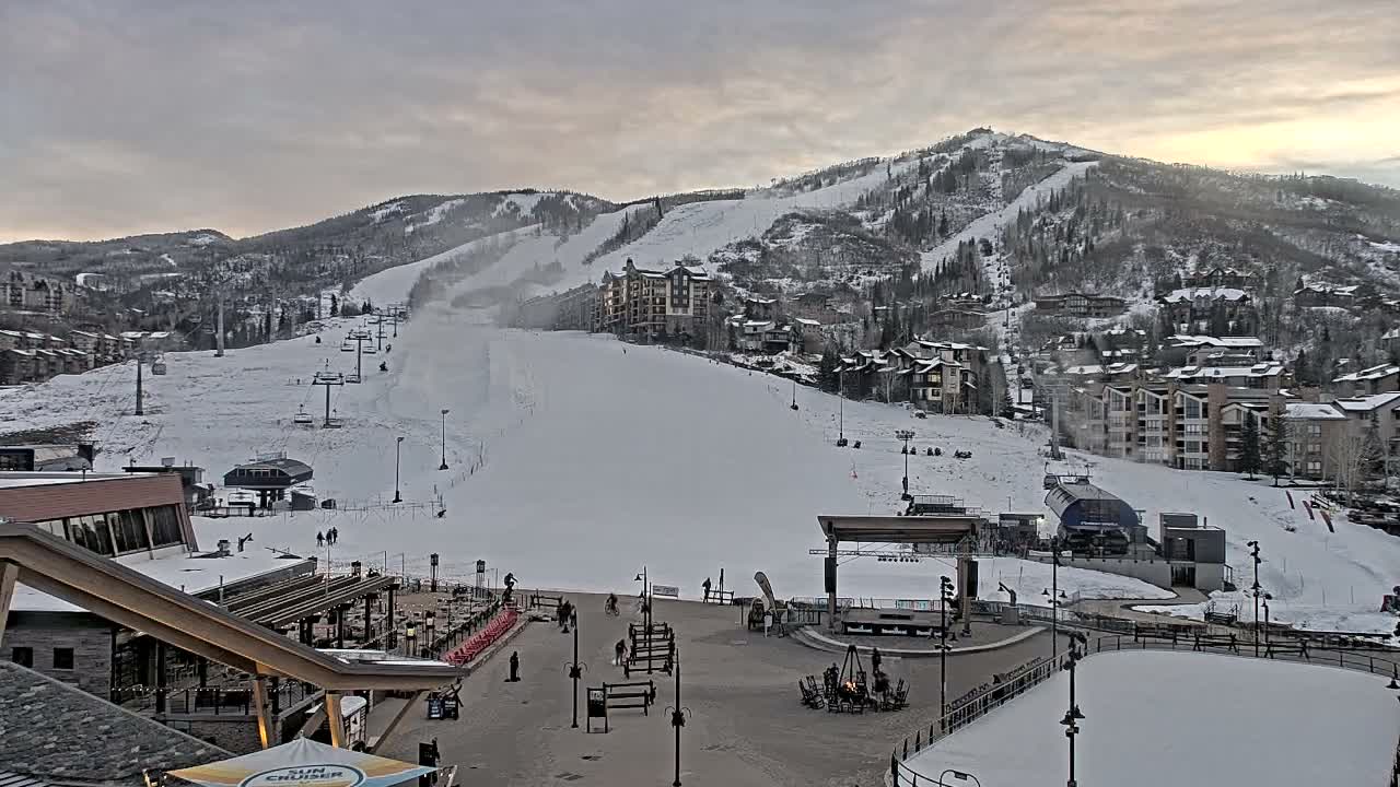 A brightly lit, snow-covered mountain ski resort village is captured at night, showcasing illuminated buildings, ski slopes with lifts, an outdoor dining area, and an ice rink on a cold, dark evening.