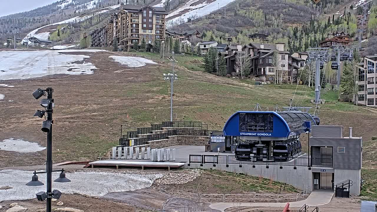 A blue gondola station sits at the base of a grassy hill dotted with buildings, with patches of melting snow visible on the hillside under a mostly clear sky.