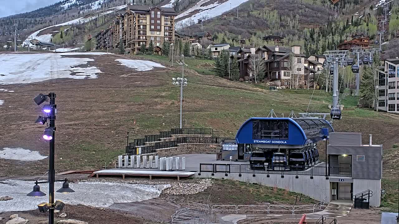A gondola station sits below a hillside of buildings and a partially snow-covered ski slope on a cloudy day.