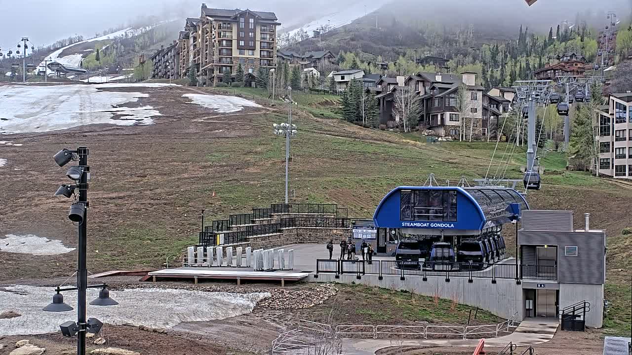 A gondola lift base station with melting snow and patchy grass sits below a mountainside residential area under an overcast sky.