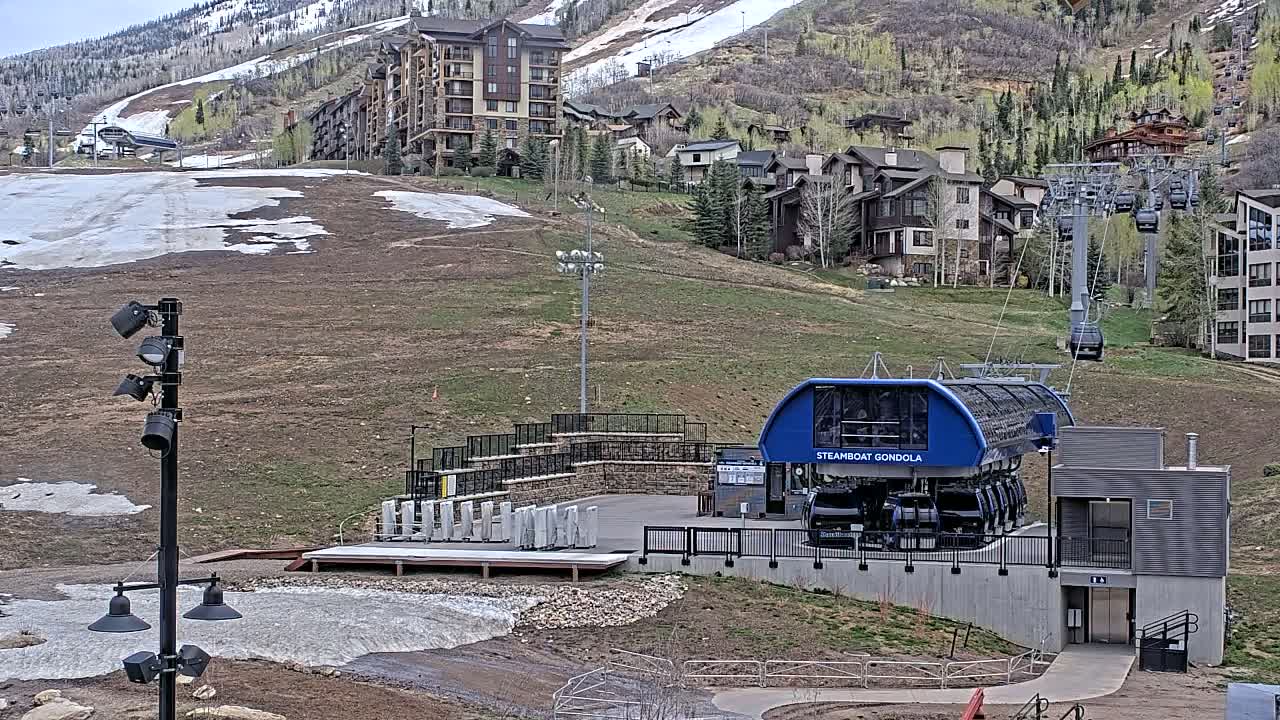 A blue gondola car sits at the base of a ski lift near residential buildings on a hillside, with patches of snow and mostly bare ground visible under a partly sunny sky.