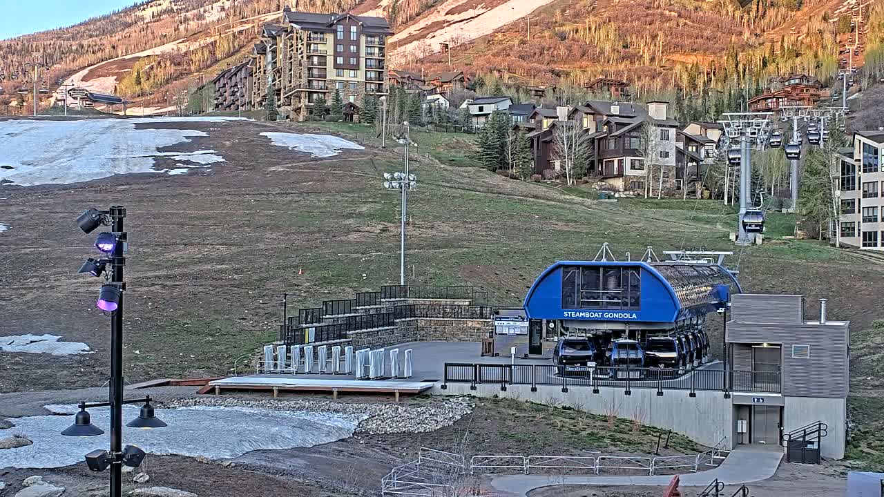 A gondola station sits at the base of a grassy hill with a partially snow-covered ski slope, overlooking a residential area and mountainside in the late afternoon.
