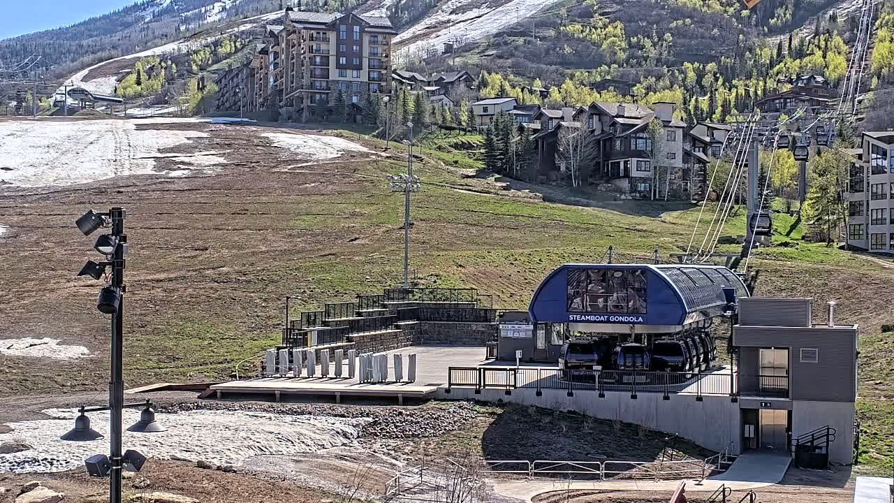 A gondola lift station sits on a grassy hillside with melting snow patches, backed by a mountain with residential buildings and partially snow-covered slopes under a sunny sky.