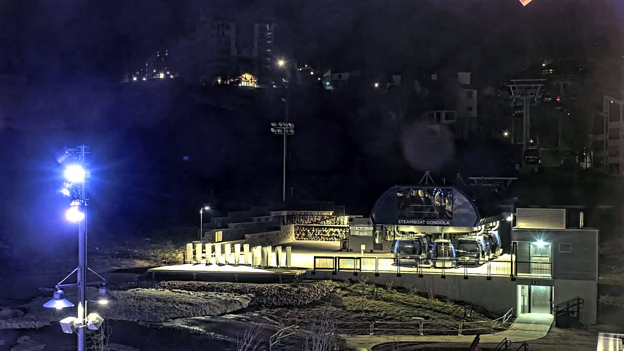 At night, a gondola station with several gondolas sits below a hill dotted with buildings and lit by streetlights.